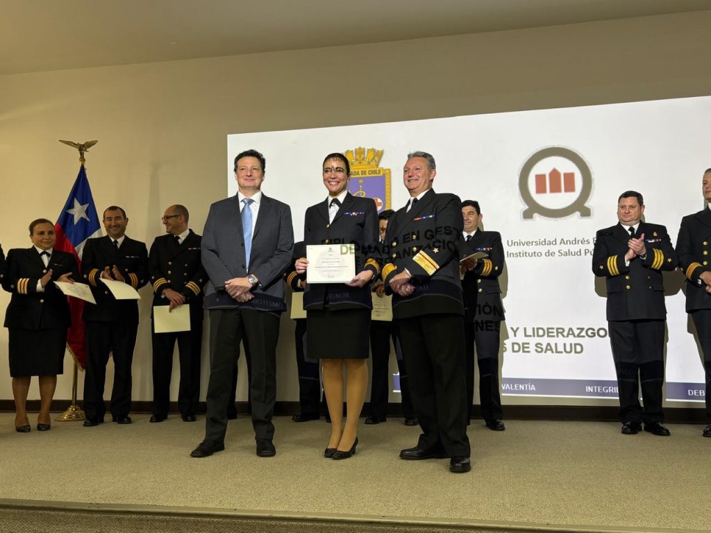 A woman in a formal uniform holds a certificate on a stage, flanked by two men in suits; other officials applaud behind them.