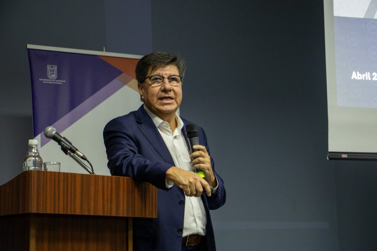 Male presenter in a dark blue suit speaks into a handheld microphone at a wooden podium; a banner and projection screen appear behind him in a conference room.