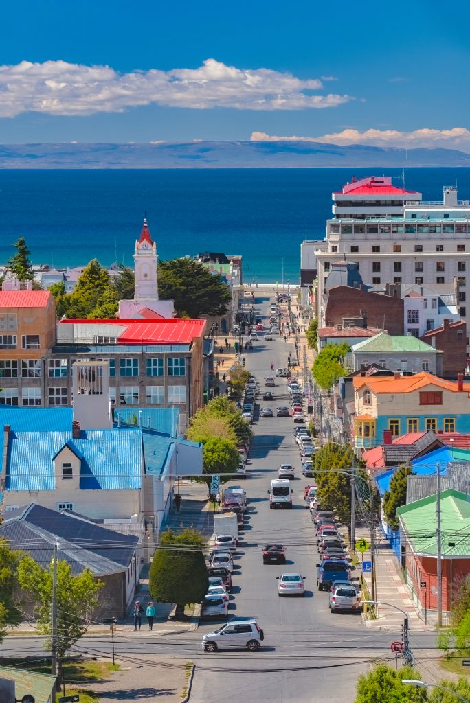 Downtown street lined with colorful buildings toward a blue sea and distant hills under a clear sky.