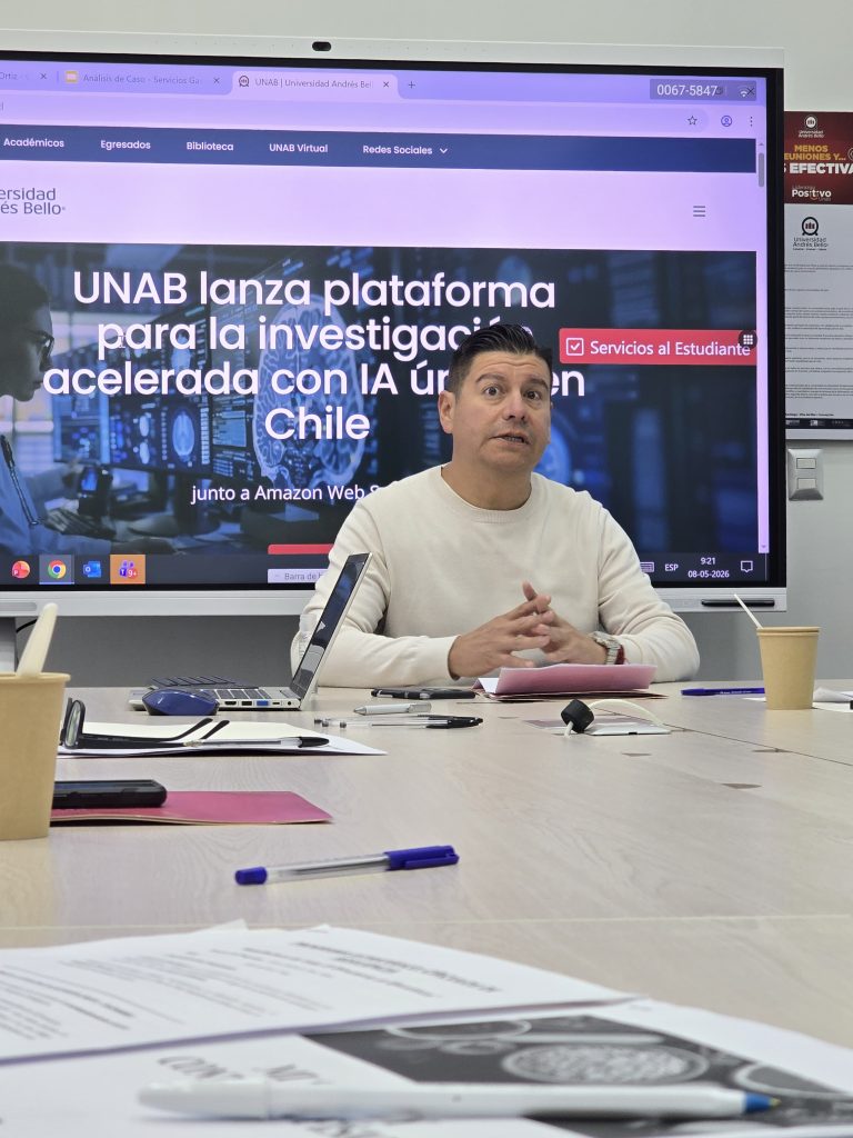 Man in a white sweater speaks at a conference table with a large screen showing UNAB branding behind him.