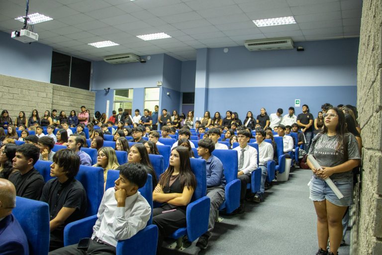 Crowded lecture hall with students seated in blue chairs; a girl with glasses stands at the right holding a rolled poster while others sit or line the back wall.