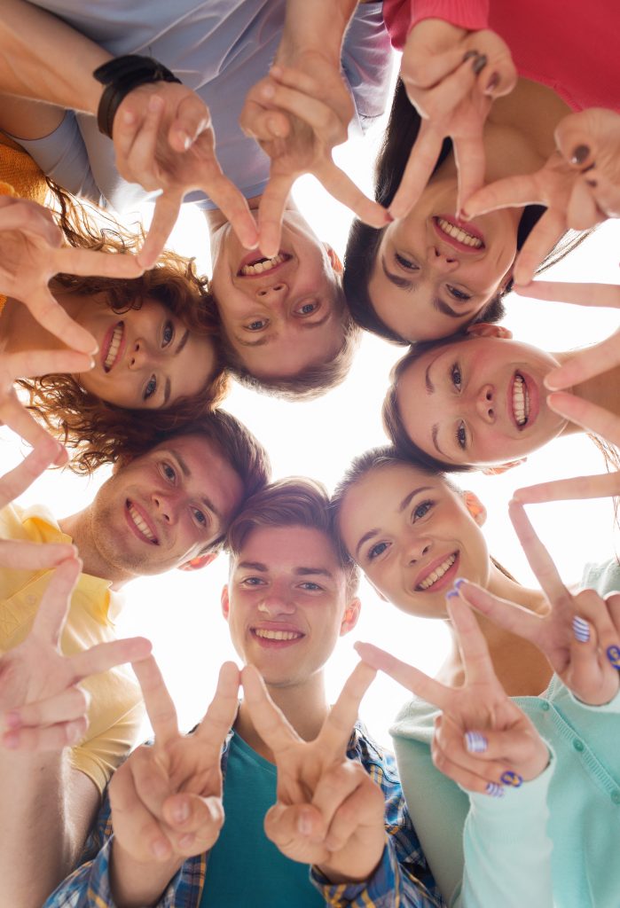 Group of smiling friends forming peace signs together, viewed from below against a bright sky