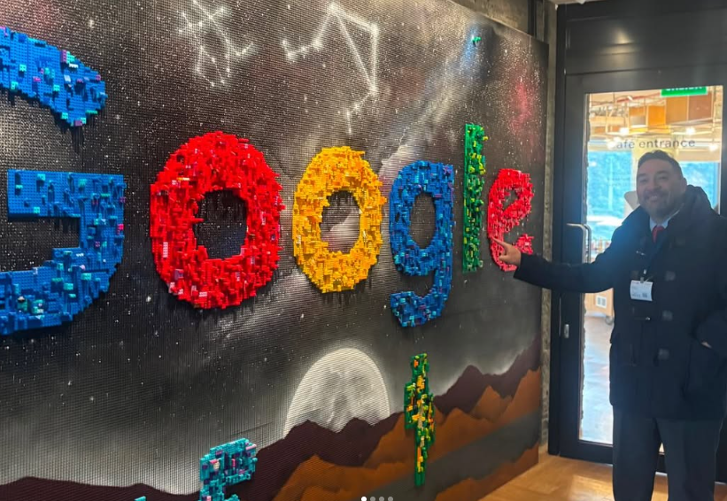 Man in a suit with a name badge stands near a cafe entrance, posing beside a wall mosaic of Google letters built from colorful blocks on a space-themed background.