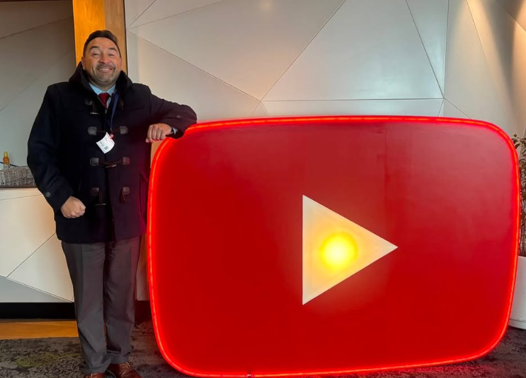 Smiling man in a dark coat stands beside a large illuminated red YouTube play button sign indoors.