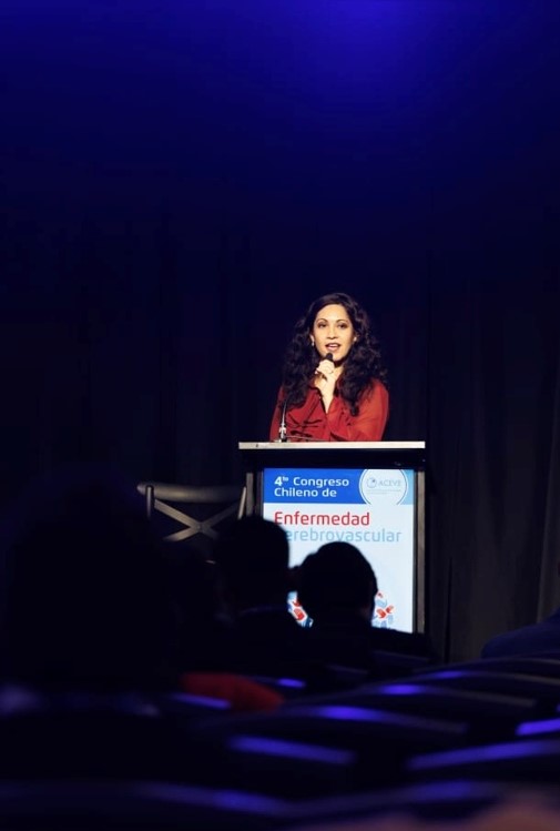 Woman at a podium on stage delivering a talk at the 4th Chilean Congress on cerebrovascular disease; audience silhouettes in foreground.