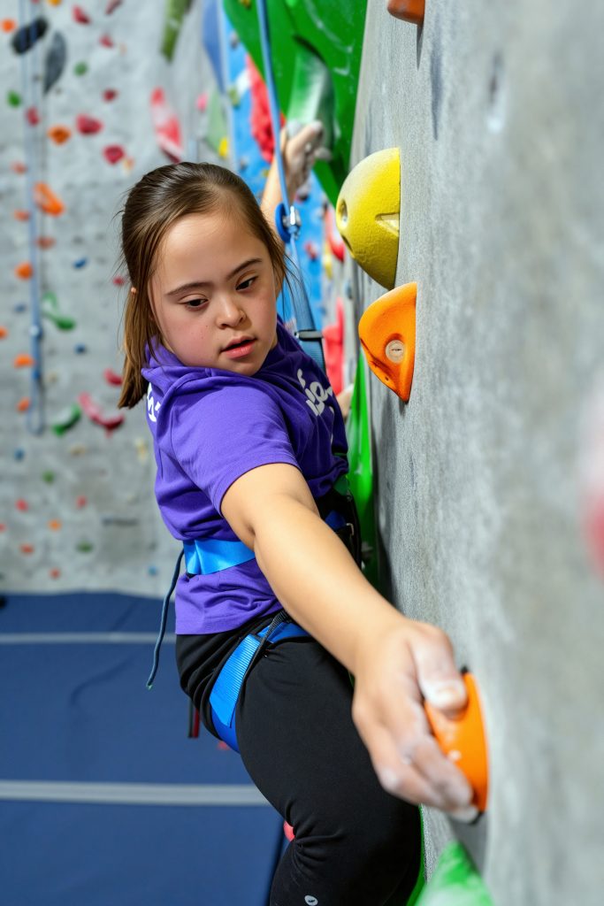 Young girl in a purple shirt wearing a climbing harness climbs an indoor rock wall, reaching for an orange hold.