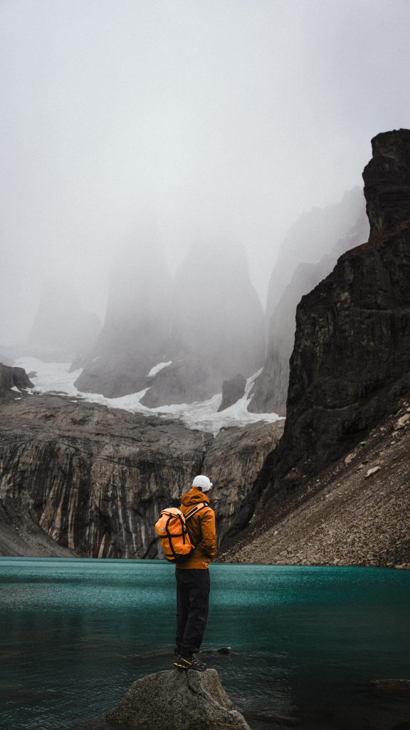 Hiker in an orange jacket and backpack standing on a rock by a turquoise glacial lake, foggy jagged mountains in the background.