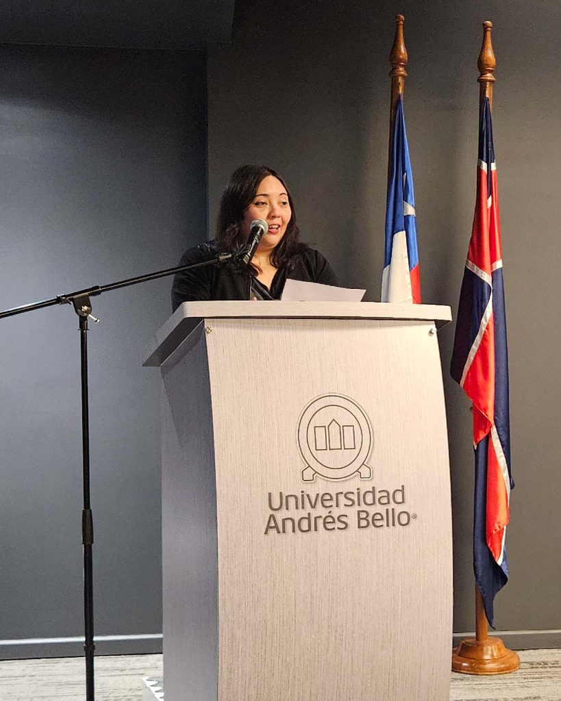 Woman speaks at a podium with a microphone, Universidad Andrés Bello emblem on the front and flags in the background.