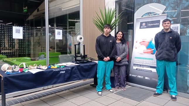 Three young people pose beside a demonstration table with equipment outdoors, a tall plant and a promotional banner in the background.