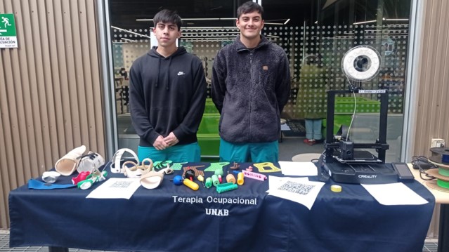 Two young men stand behind a table displaying occupational therapy tools and devices, with a sign reading Terapia Ocupacional UNAB.