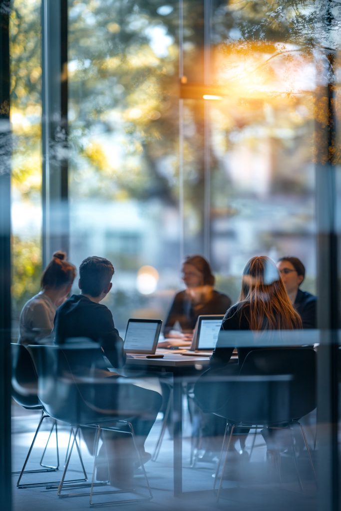 People seated around a conference table with laptops, seen through a glass-walled meeting room with sunlight.