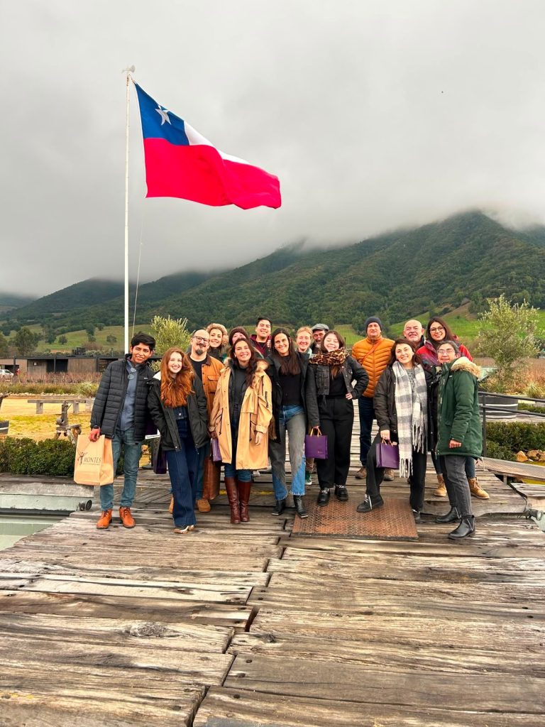 Group of about 20 people posing on a wooden dock with a large Chilean flag waving, mountains and cloudy sky in the background.