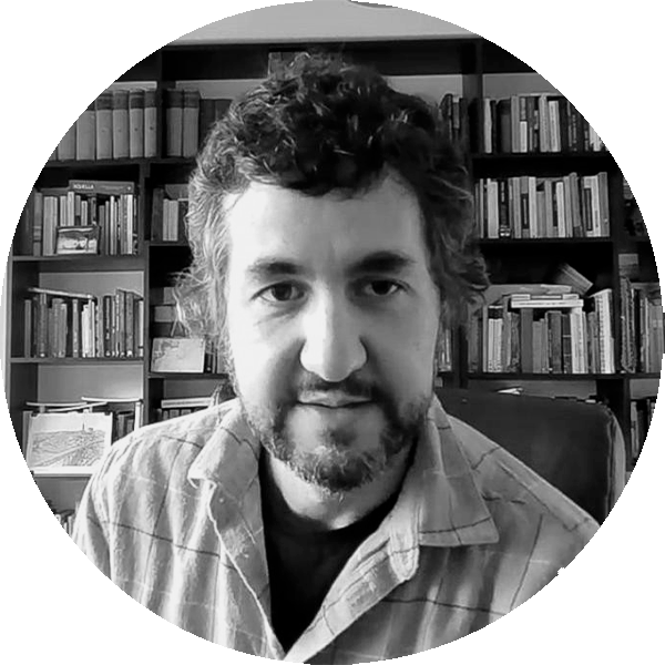 Black-and-white portrait of a man with dark hair and a beard, wearing a plaid shirt, in a room lined with books.